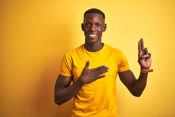Молодой африканский американец в обычной футболке, стоящей на изолированном желтом фоне, улыбаясь, ругаясь рукой на груди и пальцах вверх, давая клятву верности
 - Фото, изображение