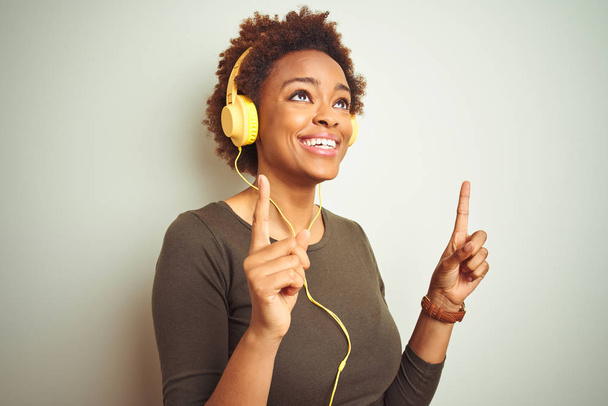 Kulaklık takan Afrikalı Amerikalı bir kadın izole edilmiş arka planda müzik dinlerken şaşkın şaşkın yukarı bakıyor ve parmaklarıyla işaret ediyor ve kollarını kaldırıyordu.. - Fotoğraf, Görsel