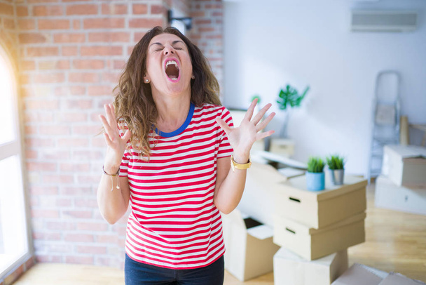 Middle age senior woman moving to a new house packing cardboard boxes crazy and mad shouting and yelling with aggressive expression and arms raised. Frustration concept. - Фото, зображення