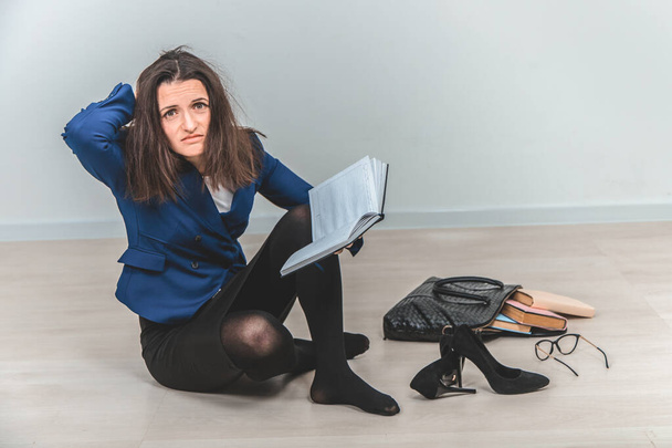 Full-length young teacher with tousled hair, sitting on the floor in complete mess around her, keeping hand on the head, looking at the camera with suffering eyes. - Foto, imagen