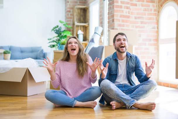 Young beautiful couple moving to a new house sitting on the floor crazy and mad shouting and yelling with aggressive expression and arms raised. Frustration concept. - Photo, Image