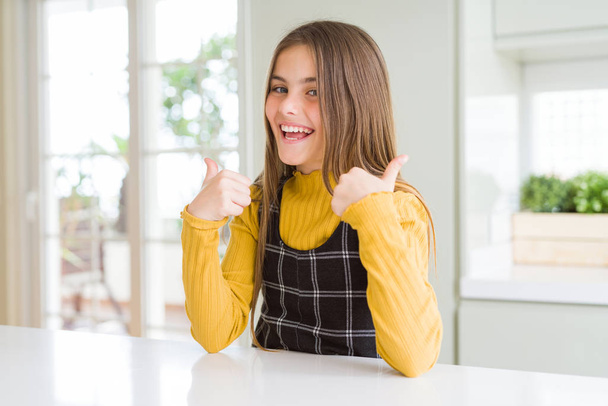 Young beautiful blonde kid girl wearing casual yellow sweater at home success sign doing positive gesture with hand, thumbs up smiling and happy. Cheerful expression and winner gesture. - Fotografie, Obrázek