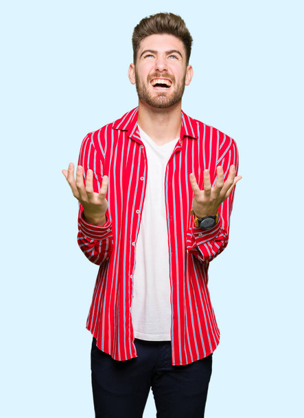 Homem bonito jovem vestindo camisa vermelha louco e louco gritando e gritando com expressão agressiva e braços levantados. Conceito de frustração
. - Foto, Imagem