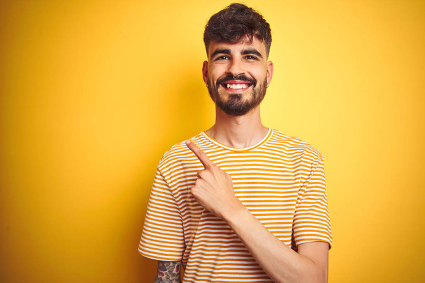 Молодой человек с татуировкой в полосатой футболке стоит на изолированном желтом фоне веселый с улыбкой на лице указывая рукой и пальцем в сторону со счастливым и естественным выражением лица
 - Фото, изображение