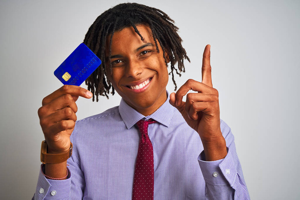 Афро-американский бизнесмен с дредами, держащий кредитную карту на изолированном белом фоне, удивленный идеей или вопросом, указывая пальцем со счастливым лицом, номер один
 - Фото, изображение
