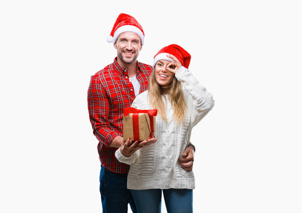 Молода пара закохана в різдвяний капелюх і тримає подарунок над ізольованим тлом з щасливим обличчям, посміхаючись, роблячи знак з рукою на око, дивлячись через пальці
 - Фото, зображення