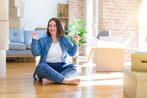 Young plus size woman sitting on the floor around cardboard boxes moving to a new home looking confident with smile on face, pointing oneself with fingers proud and happy. - Photo, Image