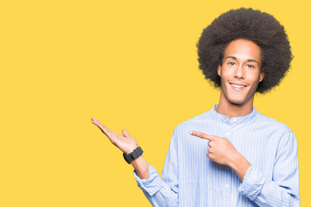 Jeune homme afro-américain aux cheveux afro étonné et souriant à la caméra tout en présentant avec la main et pointant du doigt
. - Photo, image