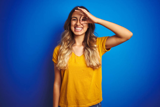 Sarı tişörtlü genç bir kadın mavi izole edilmiş arka planda el hareketleriyle gülümsüyor ve mutlu yüzüyle parmaklarının arasından bakıyor.. - Fotoğraf, Görsel