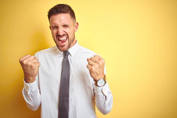 Young handsome business man wearing elegant white shirt over yellow isolated background very happy and excited doing winner gesture with arms raised, smiling and screaming for success. Celebration concept. - Valokuva, kuva