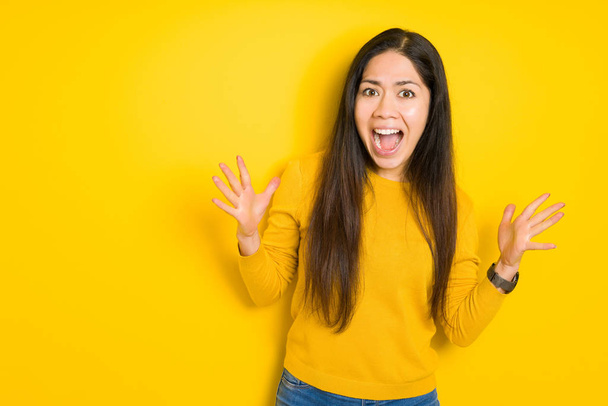 Beautiful brunette woman over yellow isolated background celebrating crazy and amazed for success with arms raised and open eyes screaming excited. Winner concept - Photo, Image