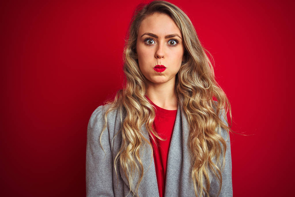 Jonge mooie zakenvrouw draagt elegante jas staande over rood geïsoleerde achtergrond puffen wangen met grappig gezicht. Mond opgeblazen met lucht, gekke uitdrukking. - Foto, afbeelding