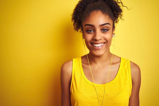 afrikanisch-amerikanische Frau, die Musik über Kopfhörer über isolierten gelben Hintergrund mit einem fröhlichen und kühlen Lächeln im Gesicht hört. Glück gehabt. - Foto, Bild