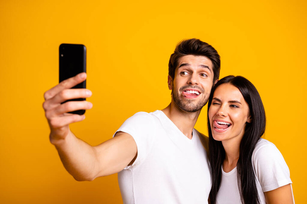Photo of two people holding telephone taking selfies sticking tongues out of mouth playful mood wear casual clothes isolated yellow color background - Photo, image