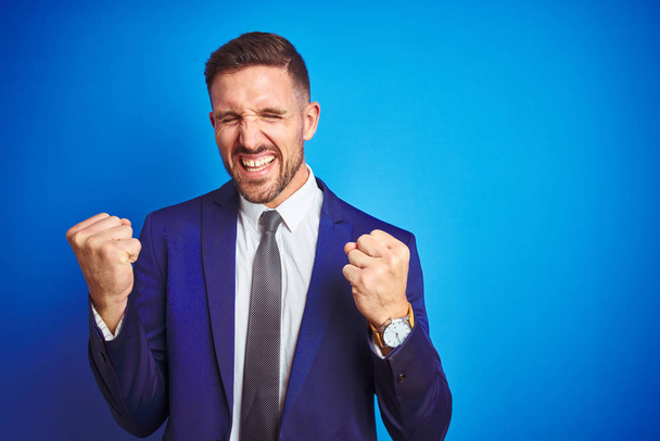 Young handsome business man over blue isolated background very happy and excited doing winner gesture with arms raised, smiling and screaming for success. Celebration concept. - Photo, Image