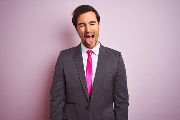 Young handsome businessman wearing suit and tie standing over isolated pink background sticking tongue out happy with funny expression. Emotion concept. - Photo, Image