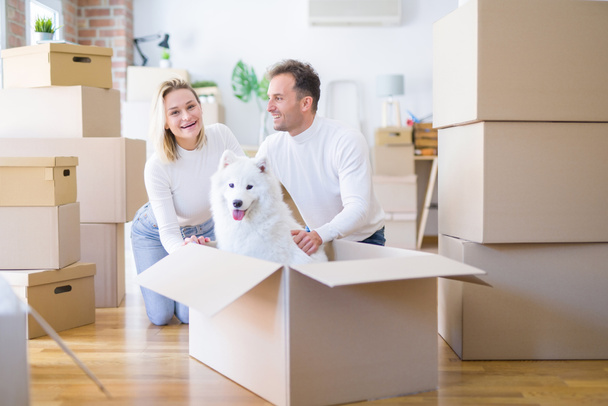 Young beautiful couple with dog sitting on the floor at new home around cardboard boxes - Fotografie, Obrázek