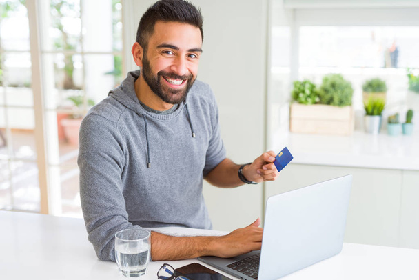 Knappe man glimlachend met creditcard als betaling metod bij het winkelen online met behulp van laptop - Foto, afbeelding