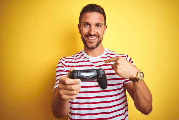 Fiatal jóképű férfi játszik videojátékok segítségével joystick játék pad sárga háttér meglepetés arc ujjal mutogatott magára - Fotó, kép