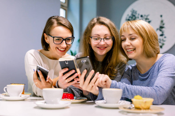 Group of three young women smiling and showing the pictures on the smartphones each other, sitting in restaurant, drinking coffee and eating desserts - Fotó, kép