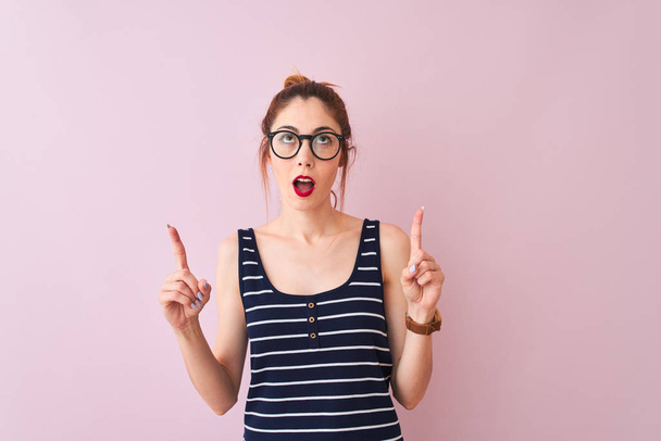 Redhead woman with pigtail wearing striped t-shirt standing over isolated pink background amazed and surprised looking up and pointing with fingers and raised arms. - Photo, Image