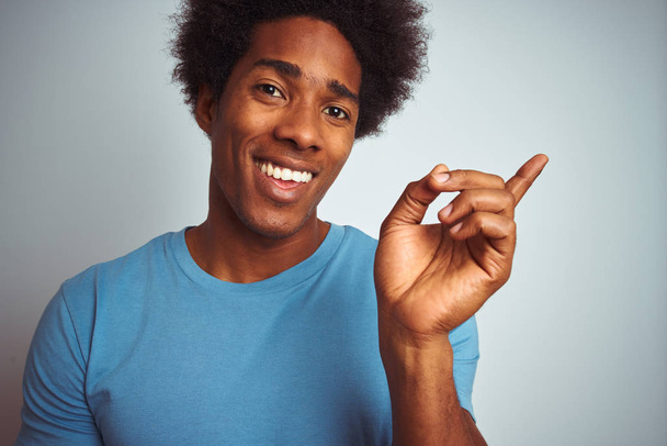 Afričan Američan s afro vlasy na sobě modré tričko stojící nad izolovaným bílým pozadím velmi šťastný ukazující rukou a prstem na stranu - Fotografie, Obrázek