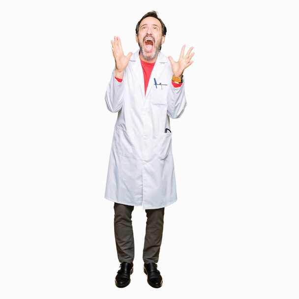 Middle age therapist wearing white coat crazy and mad shouting and yelling with aggressive expression and arms raised. Frustration concept. - Фото, изображение