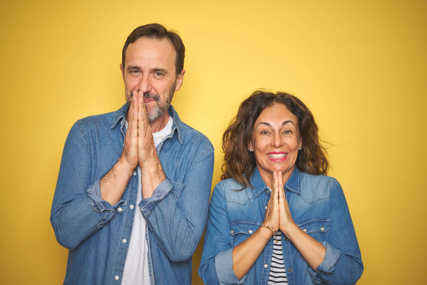 Krásný pár středního věku spolu nosí džínové košile přes izolované žluté pozadí modlí s rukama dohromady prosí o odpuštění s úsměvem sebevědomí. - Fotografie, Obrázek