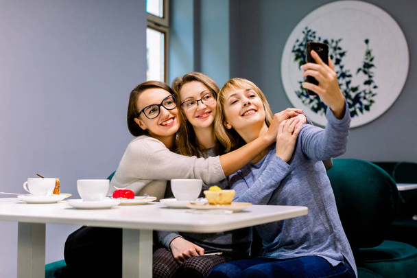 Three beautiful young women enjoying coffee and cake together in a cafe sitting at a table laughing and making selfie photo with happy smiles - Valokuva, kuva