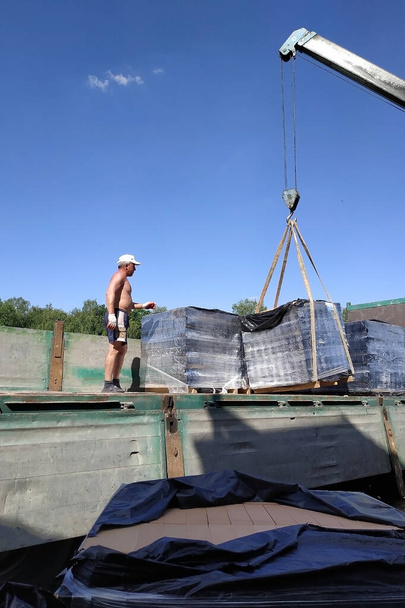 移動式クレーンの助けを借りて建設のためのフロントレンガでパレットをアンロード村の労働者. - 写真・画像