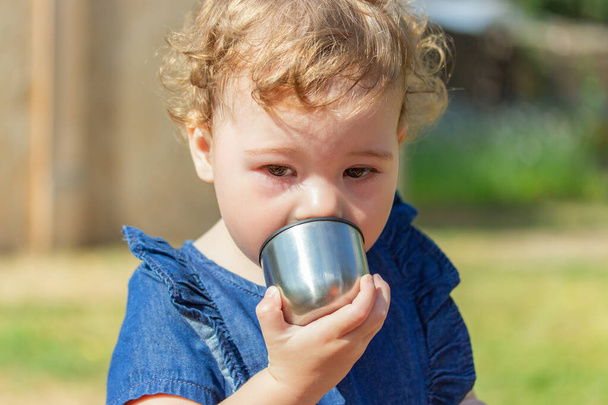 Bambina in un abito di jeans beve da una tazza di metallo, ritratto di un primo piano bambino di 1 anno. Ragazza caucasica tiene una tazza da un thermos cromato
 - Foto, immagini