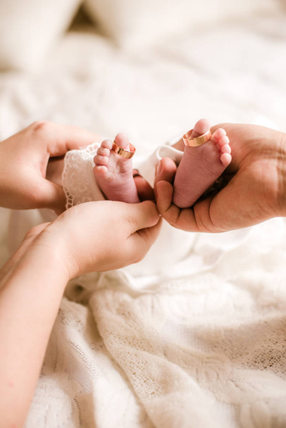 Mom's and father hands are holding little cute legs of a newborn baby at home on a white bed. Happy motherhood - Photo, Image
