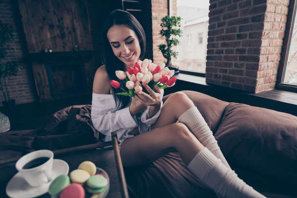 Portrait of her she nice attractive charming pretty cheerful dreamy girl sitting on bed holding in hands enjoying smelling fresh tulips at industrial brick wood loft modern interior style house - Foto, afbeelding