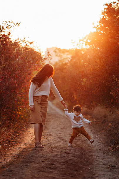 Mały chłopiec i jego matka spacerują w jesiennym parku. Piękny zachód słońca, czerwone liście - Zdjęcie, obraz