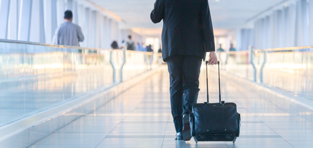 Businessman walking and wheeling a trolley suitcase at the lobby, talking on a mobile phone. Business travel concept. - Photo, Image