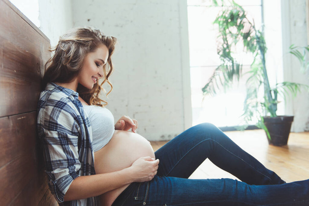 Happy pregnant woman sitting on the floor and touching her belly at home - Photo, Image