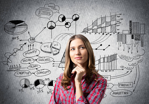 Brown haired woman looks pensive upwards and thinking something. Financial charts and business doodles on grey wall. Puzzled girl has serious facial expression. Woman wears red checkered shirt - 写真・画像