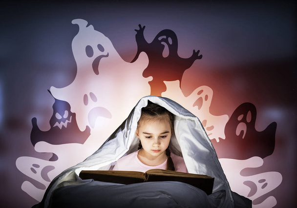 Entuzjastyczna dziewczynka czytanie książki w łóżku po snem. Kid w piżamie z latarką ukrywając się pod kocem. Przerażona dziewczyna i wymyślonych potworach na głębokim niebieskim tle. Czytanie bajek dla dzieci. - Zdjęcie, obraz