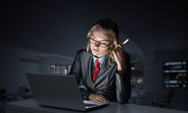 Attractive young woman in glasses sitting at desk and working at laptop. Caucasian businesswoman working overtime in office to finish project within deadline. Career building and business development - Foto, Imagen