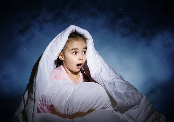 Surprised girl with pillow hiding under blanket. Amazed kid lying in his bed at home. Fear of the dark. Covered kid can not sleep at night. Portrait of girl in pajamas on background of deep blue sky - Valokuva, kuva