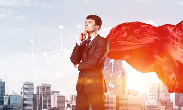 Young pensive businessman wearing red cape against modern city background - 写真・画像