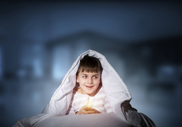 Happy child with flashlight hiding under blanket. Smiling kid lying in bed at home. Funny game at bedtime. Young boy can not sleep at night. Cheerful boy in pajamas on deep blue blurred background. - Foto, Imagen