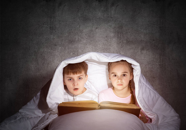 Frightened girl and boy looking at each other under blanket. Children reading magic stories in bed before going to sleep. Young sister and brother in pajamas together on background of grey wall. - Photo, Image