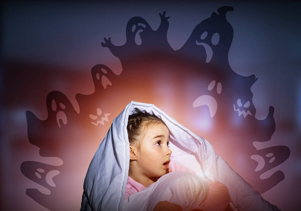 Возбужденная девушка прячется под одеялом от воображаемых призраков. Испуганный ребенок сидит дома в своей постели. Маленькая девочка боится выдуманных монстров. Покрытый пижамой ребенок не спит по ночам. Хэллоуин
. - Фото, изображение