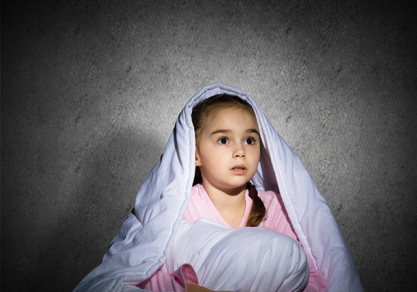 Frightened girl hiding under blanket. Scared kid lying in his bed at home. Fear of the dark. Beautiful little girl can not sleep at night. Portrait of child in pajamas on background of grey wall. - Fotografie, Obrázek