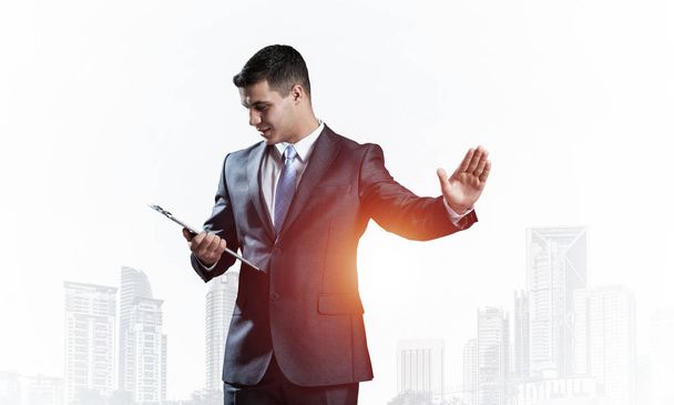 Young elegant businessman standing and looking into documents. Caucasian man in business suit and tie on background of modern cityscape. Successful business person with raised hand and open palm. - Fotografie, Obrázek