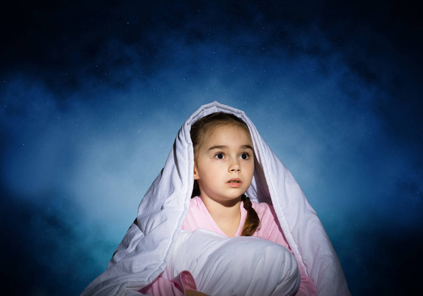 Scared girl with pillow hiding under blanket. Startled kid lying in his bed at home. Little girl afraid of dark. Covered child not sleep at night. Girl in pajamas on background of deep blue sky. - Photo, Image