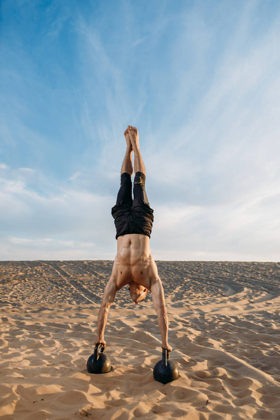 Male athlete doing exercises with two kettlebells stands on hands in desert at sunny day. Strong motivation in sport, strength outdoor training - Fotografie, Obrázek