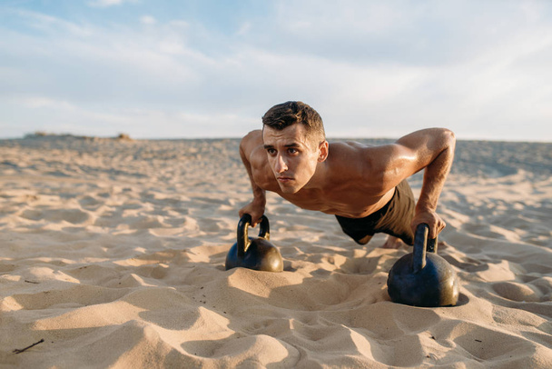 Mannelijke atleet doet push-up oefeningen met twee kettlebells in de woestijn op zonnige dag. Sterke motivatie in de sport, krachttraining buitenshuis - Foto, afbeelding