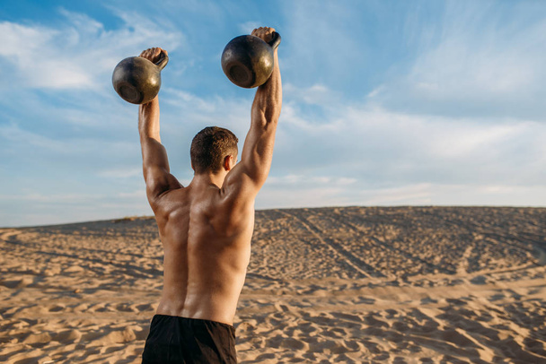 Άντρας αθλητής κάνει ασκήσεις με δύο κέτλμπελ στην έρημο την ηλιόλουστη μέρα. Ισχυρό κίνητρο στον αθλητισμό, δύναμη υπαίθρια εκπαίδευση - Φωτογραφία, εικόνα